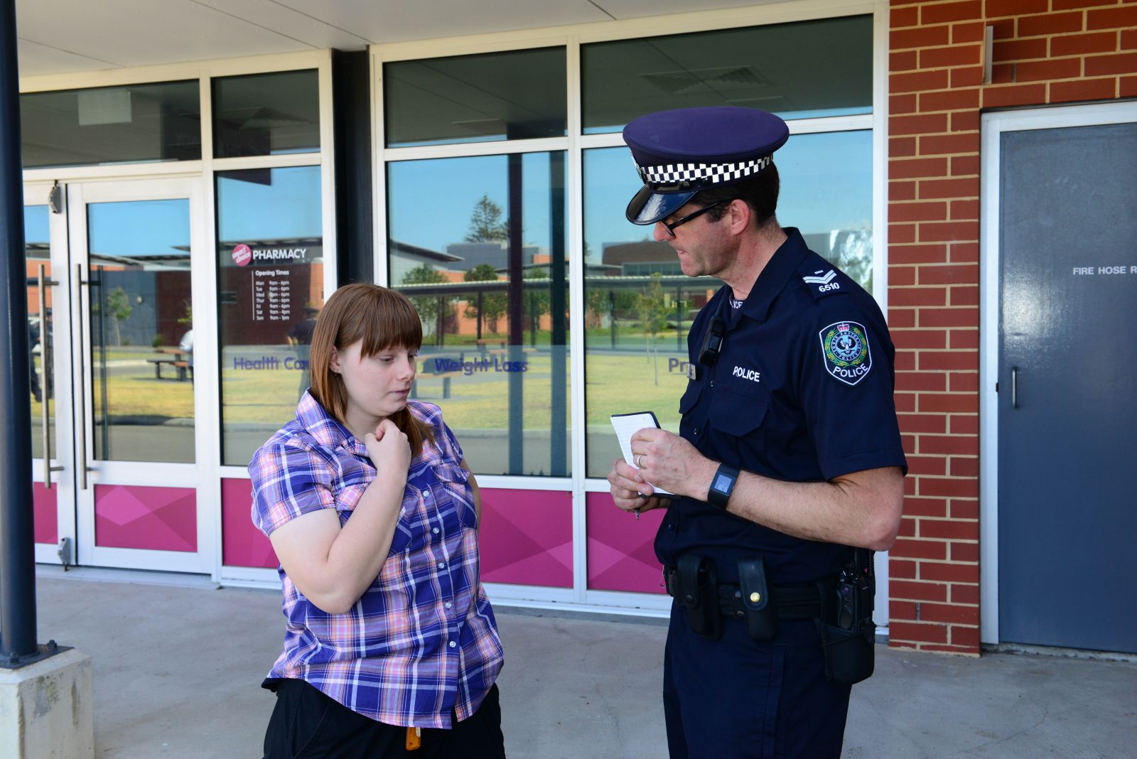 A woman talks to a police officer outside of a chemist. The police officer is writing down what she says in a notebook.
